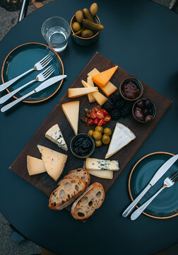  gourmet cheese platter with assorted colorful cheeses, seeds, jam, blackberries, olives, pickled vegetables, rustic bread and elegant cutlery, arranged on a dark modern table at an outdoor caf&eacute;. 