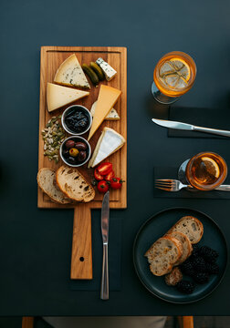  gourmet cheese platter with assorted colorful cheeses, seeds, jam, blackberries, olives, pickled vegetables, rustic bread and elegant cutlery, arranged on a dark modern table at an outdoor caf&eacute;. 