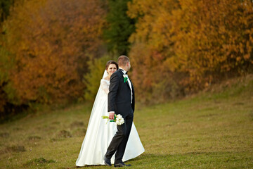 A bride and groom are standing in a field with a beautiful fall backdrop. The bride is holding a...
