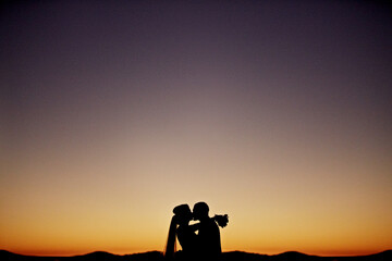 A couple is kissing in front of a beautiful sunset. The sky is a deep purple color, and the...