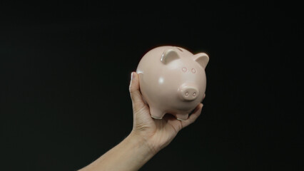 Hand holding pink piggy bank against black background, representing savings and financial security,...