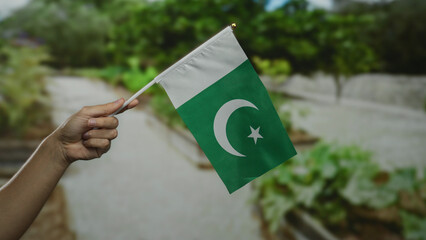 Hand holding pakistan flag outdoors in a city street setting with garden background, signifying...