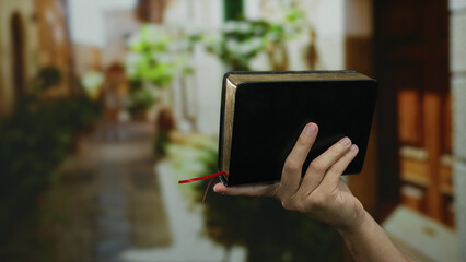 Man holding a book on a sunny street, showcasing the serene atmosphere of a town with blurred green plants.