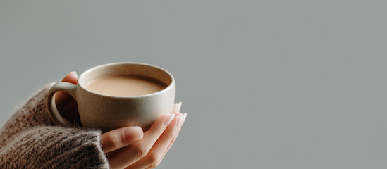 Woman in a cozy sweater holds a warm ceramic mug of coffee against a plain gray background with copy space banner.