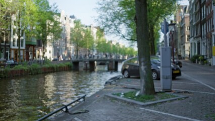 Fototapeta premium Blurred view of amsterdam canal with defocused buildings, tree, cars, and bridge on a sunny day showcasing the serene urban landscape.