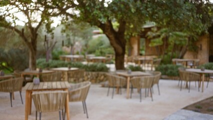 Blurred view of outdoor mediterranean restaurant terrace with wooden chairs and tables under trees offering a serene dining experience in a defocused setting.