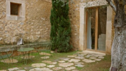 Fototapeta premium Blurred view of an old mediterranean house exterior with stone walls, green patio furniture, and a tree, capturing a serene outdoors ambiance.
