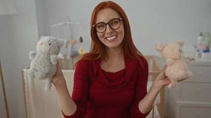 Woman with red hair smiles holding cute plush toys in cozy bedroom with a crib and dresser in the...
