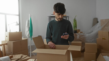 Young man unpacking boxes in a new apartment, holding a smartphone, surrounded by moving supplies,...