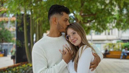 Obraz premium Couple embracing warmly in an urban park setting with lush green trees, conveying love and connection between a man and woman in a serene outdoor environment.