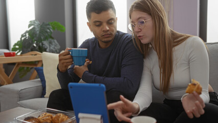 Couple sitting indoors on sofa with coffee and pastries discussing over a tablet, man and woman in living room setting having casual conversation and interaction