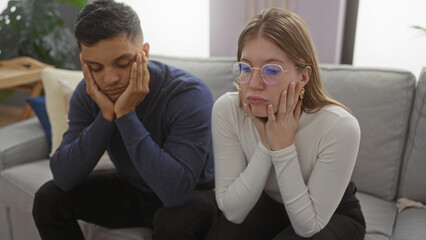 Couple sitting on couch indoor looking pensive in living room, showcasing relationship dynamics in...