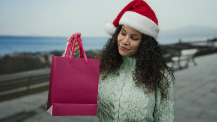 Woman in santa hat holds pink bag by the sea on a promenade showcasing christmas spirit in an...