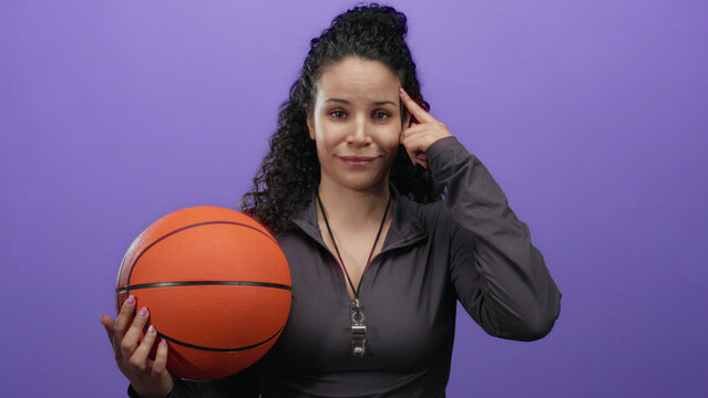 Woman holding basketball in front of a purple wall while pointing at her temple, suggesting a thoughtful pose in a sports context, wears casual sports attire with a whistle.