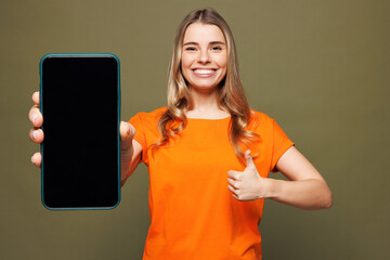 Young woman wears orange t-shirt casual clothes hold in hand use mobile cell phone with blank screen workspace area show thumb up isolated on plain pastel green background studio. Lifestyle concept.
