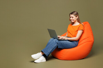 Full body young smiling IT woman wears orange t-shirt casual clothes sit in bag chair hold use work...