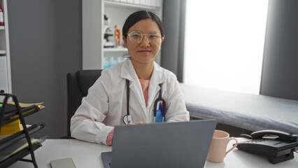 Young woman in a medical uniform with a stethoscope sits at a desk in a modern clinic room,...