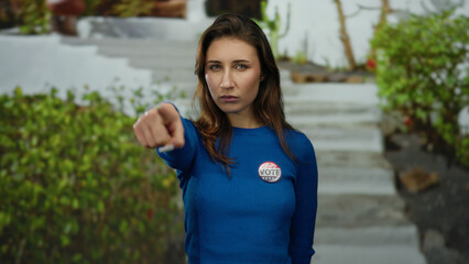 Woman in a blue sweater with a 'vote' badge points assertively outdoors on a city street,...