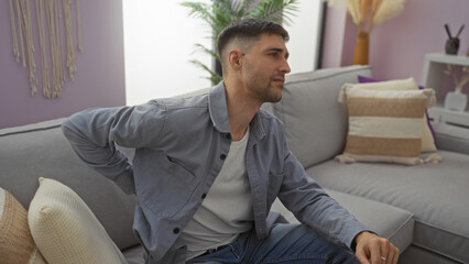 Young man with back pain sitting on a couch in a modern living room, expressing discomfort,...