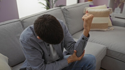 Young man sitting on a couch in a modern living room holding his elbow indicating discomfort, wearing casual clothes, in a relaxed domestic setting, showcasing a home environment.