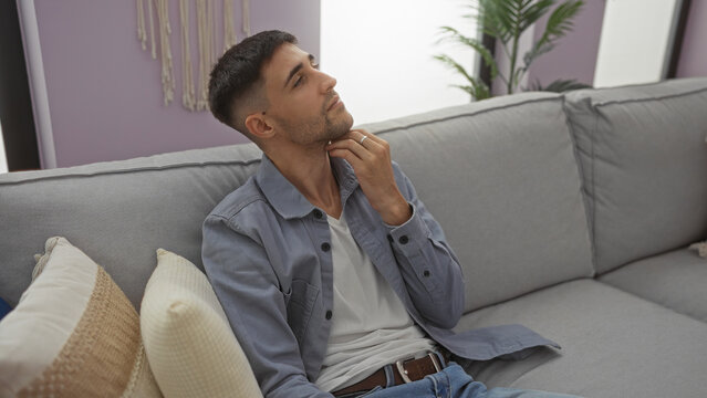 Young man sitting thoughtfully on a comfortable sofa in a stylish living room, with hispanic features and a casual outfit, surrounded by cozy home decor. - Powered by Adobe