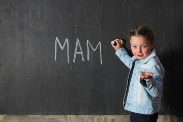 A little girl writes MAM with chalk on a chalkboard