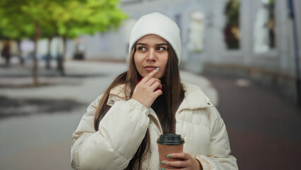 Woman pondering with coffee cup outdoors wearing beanie showing thoughtful expression in city...