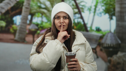 Woman wearing winter jacket holding mate drink in park making silence gesture with finger to mouth...
