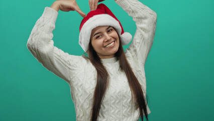 Hispanic woman wearing santa hat making heart gesture with hands over head isolated on green background smiling joyfully suggesting christmas spirit and happiness.