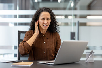 A woman with curly hair is in pain, holding her ear while working on a laptop in an office setting.