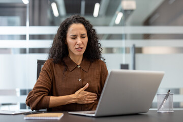 A woman experiencing chest pain while working at a laptop in an office setting, possibly indicating...