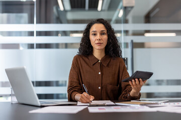 A businesswoman with curly hair works on documents in an office setting, using a calculator and...