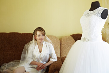 A bride sits on a couch with a wedding dress in front of her. The dress is white and has lace...