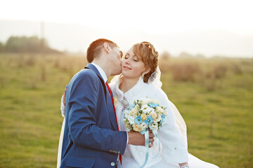 A bride and groom kiss in a field. The bride is wearing a white dress and a veil. The groom is...