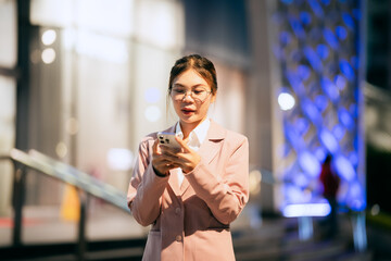 A confident young businesswoman stands outdoors at night in a modern city, smiling while using her smartphone, reflecting success, style, ambition, and digital connectivity.