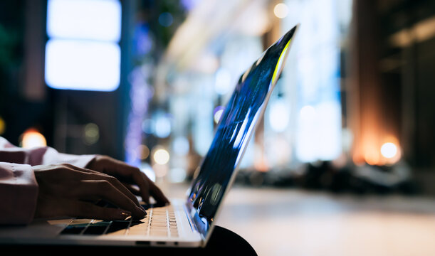 Close-up of a person typing on a laptop at night with blurred lights in the background, symbolizing technology, remote work, focus, and modern digital life.
