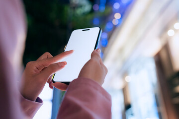 A close-up of a woman using a smartphone with a blank white screen at night. Technology, communication, and digital interaction in a modern urban setting.