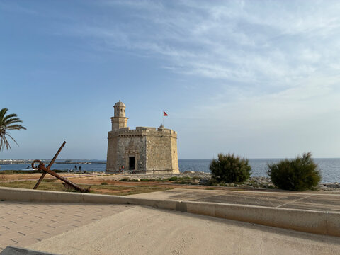 Historic stone watchtower on the coast of Menorca, with a weathered anchor in the foreground, palm trees, Mediterranean Sea a clear blue sky ideal for travel, history, and coastal themes.