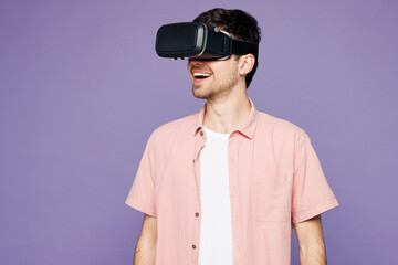 Young smiling happy cheerful fun cool man wears pink shirt t-shirt casual clothes watching in vr headset pc gadget isolated on plain pastel light purple background studio portrait. Lifestyle concept.