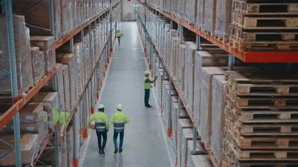 Workers in protective helmets moving between massive storage shelves filled with boxes. Two employees carrying package. Employees walking or inspecting stacked inventory along warehouse aisle. - Powered by Adobe