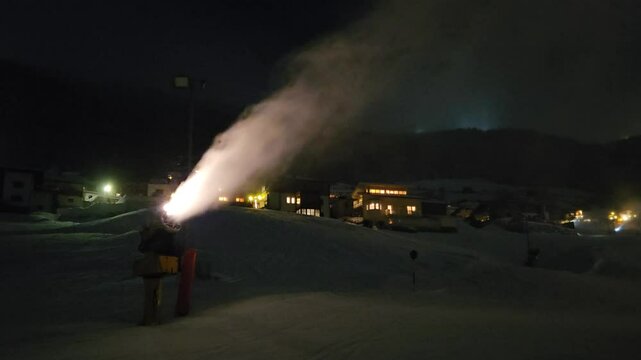 blowing snow with a snow blower in front of a building. The scene is dark, and the frozen water is falling heavily on ski resort
