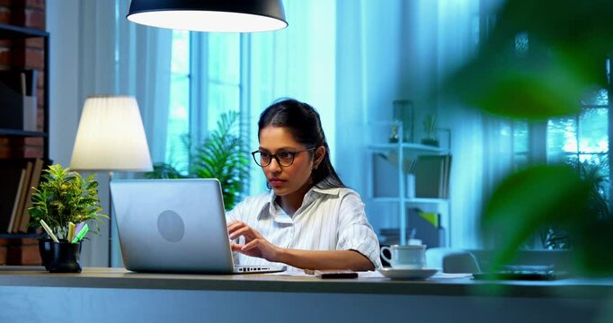 Indian businesswoman drafting email on laptop at desk in modern cozy office, Asian young female corporate executive typing an important message while thinking working remotely on wireless computer