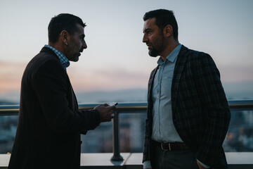 Two professionals engaged in a discussion while standing on a rooftop with a sunset view, promoting...