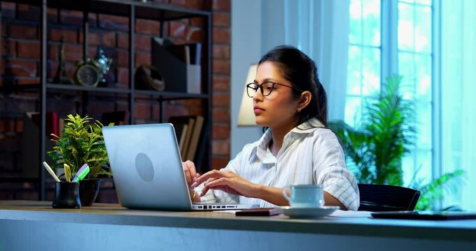 Indian businesswoman drafting email on laptop at desk in modern cozy office, Asian young female corporate executive typing an important message while thinking working remotely on wireless computer