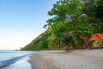 White Sand Beach at dawn: calm sea meets shadowed shore with rope swing, palm trees framing hidden bungalows in morning light. Koh Chang, Thailand.

