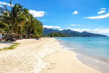 Klong Prao Beach morning: pristine white sand meets sparkling turquoise sea with swimmers, framed by palms against island mountains. Tropical paradise. Koh Chang, Thailand.

