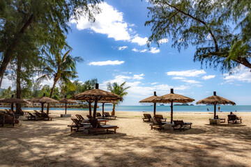 Klong Prao Beach with rustic thatched parasols shading wooden sunbeds under palms, leading to turquoise waves and island-dotted horizon. Koh Chang, Thailand.

