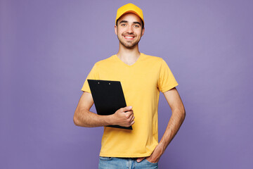 Young happy delivery guy employee man wear yellow cap t-shirt uniform workwear work as dealer courier hold clipboard with papers document isolated on plain pastel purple background. Service concept.