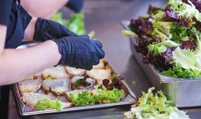 Hands in black gloves preparing healthy sandwiches with fresh lettuce in a professional kitchen. Food hygiene, catering service, or restaurant prep concept.