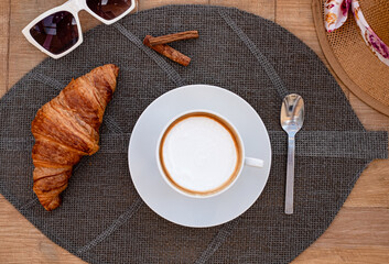 A cozy breakfast scene featuring a cup of cappuccino on a saucer, a golden croissant, cinnamon...